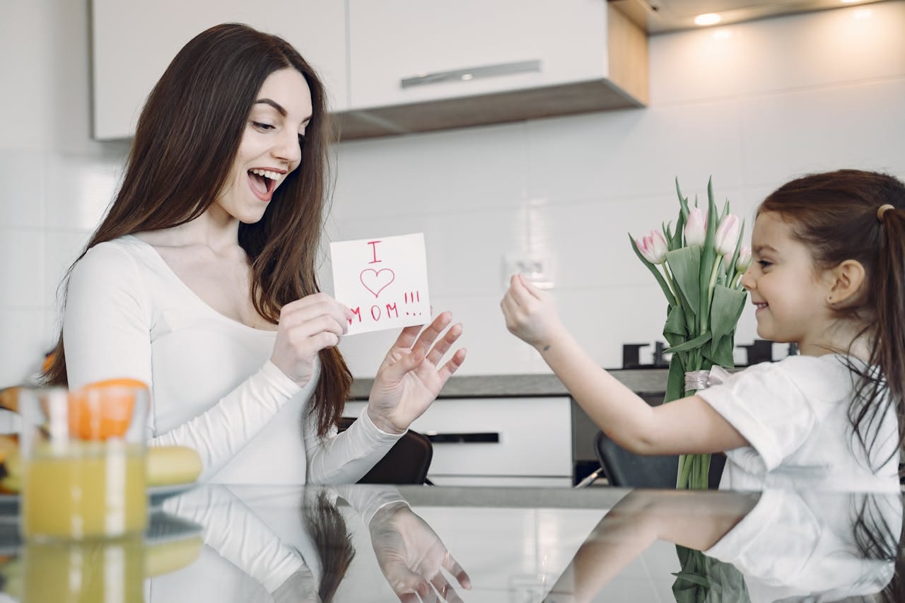 A mother and daughter sharing a joyful moment with flowers and a card in a kitchen.